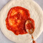 There is a large spoon spreading some pizza sauce over a no yeast sourdough pizza dough.