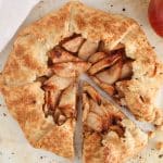 Vegan apple galette on parchment paper with a slice taken off and some fresh apples on the side.