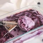 An ice cream scoop containing vegan blueberry ice cream and placed over the container with the ice cream.