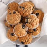 A pile of vegan banana blueberry oatmeal muffins over a white towel with fresh berries on the side.