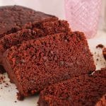 A half sliced vegan chocolate loaf cake on parchment paper with a glass of milk in the background.