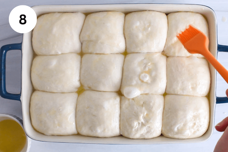 A brush is brushing melted butter over balls of dough in a baking dish.