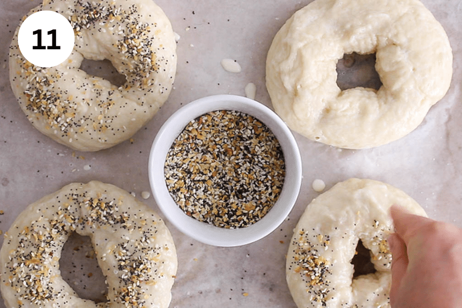 A baking sheet with brushed bagel and a hand sprinkling everything seasoning.