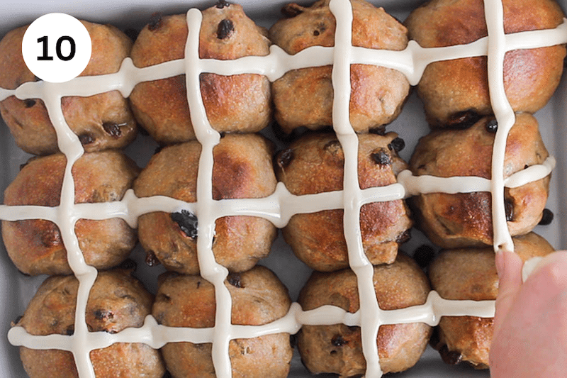 A baking dish with baked hot cross buns and a hand making X shape with frosting.