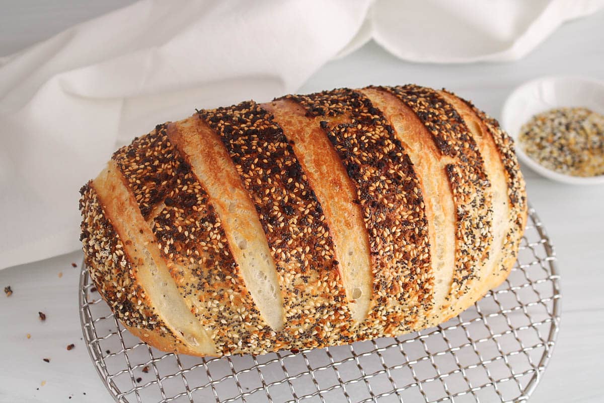A loaf of everything bagel sourdough bread on a cooling rack showing its surface covered with everything bagel seasoning.