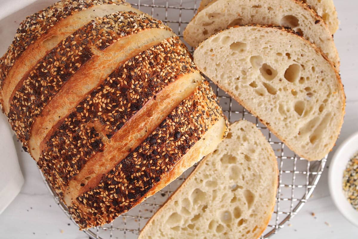 A half sliced loaf of everything bagel sourdough bread placed on a cooling rack.